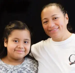 A portrait of a girl and a young woman. The girl on the left is around 10 years old and has a close-mouthed smile. She has long dark hair in a half-ponytail and wears a white and gray sweater. Next to her, the young woman has her arm around the girl's shoulders and smiles with teeth. She has dark hair pulled back and wears a white t-shirt.