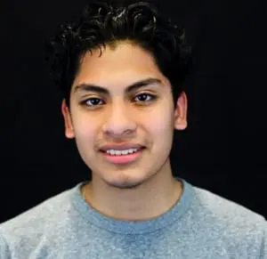 A portrait of Erik, who is a young man with a medium skin tone, short curly dark hair, and a faint moustache and goatee. He smiles with teeth and wears a gray shirt.