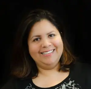 A portrait of Isela, who is a young woman with a medium skin tone and shoulder-length dark brown hair. She smiles toothily and wears a white top with a black collar and black floral embroidery.