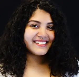 A portrait of Maria, who is a young woman with long curly dark hair and a nose ring. She smiles toothily and wears a white knit shirt.