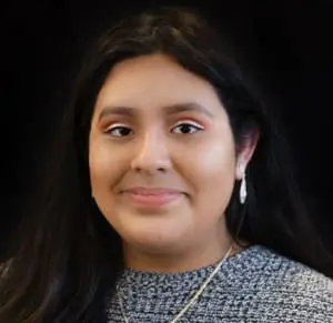 A portrait of Maria who is a young woman with a medium skin tone and a close-mouth smile. She has long, dark wavy hair, and she wears sparkling tear-drop earrings and a salt-and-pepper gray sweater.